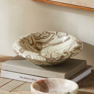 May include: A shallow ceramic bowl featuring a marbled pattern of cream and brown swirls. The bowl has a wide, rounded rim and a smooth, glossy finish. It is positioned centrally, showcasing the intricate, organic color blending throughout.