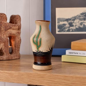 May include: A small ceramic vase with a cream-colored upper body featuring a green painted cactus design. The lower section is a dark brown band with black painted desert plant silhouettes. The vase has a narrow neck and a flared rim.
