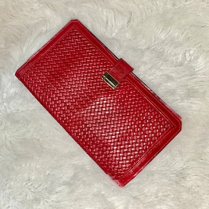 May include: A red wallet with a woven texture and a gold-colored clasp. The wallet is rectangular and has a flap closure. The background is a white, fluffy surface.
