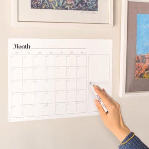Pode incluir: Um calendário mensal branco de apagar a seco com a palavra "Month" no topo. O calendário é dividido em quadrados para cada dia do mês. Uma mão segura uma caneta de apagar a seco, pronta para escrever no calendário.