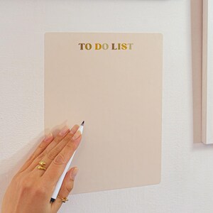 Può includere: Una lavagna beige per la lista delle cose da fare con la scritta dorata "TO DO LIST". Un pennarello bianco è tenuto vicino alla lavagna. La lavagna è montata su un muro bianco, con un'immagine incorniciata sopra.