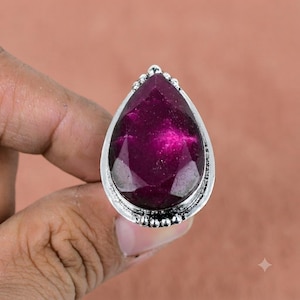 Peut inclure: Une bague en argent ornée d'une grande pierre précieuse facettée en forme de poire, d'un magenta profond. Le sertissage présente un bord perlé décoratif autour de la pierre, et l'anneau est en métal argenté poli avec une finition texturée oxydée.