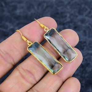 Może przedstawiać: Para złotych kolczyków z prostokątnymi, fasetowanymi, jasnozielonymi kamieniami szlachetnymi. Kolczyki mają prosty, elegancki design ze złotą oprawą i zapięciem na haczyk. Kamienie mają około 2.5 cm długości.