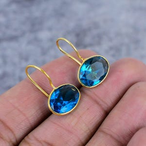 Op de afbeelding: Een paar gouden oorbellen met ovale blauwe topaas edelstenen. De oorbellen hebben een eenvoudig, elegant ontwerp.