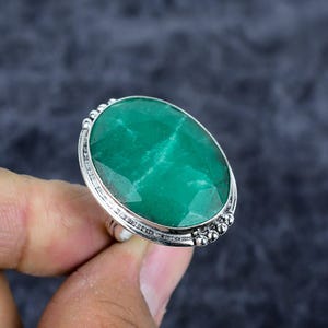 May include: A silver ring featuring a large, oval-cut, emerald-green gemstone. The gemstone has visible internal patterns and is set in a detailed silver band with small decorative beads around the edge. The ring is held up by a hand.