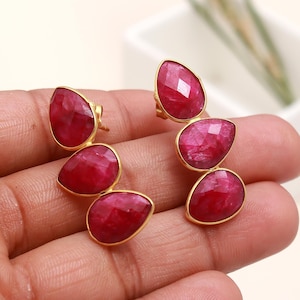 Puede incluir: Un par de pendientes dorados, cada uno con tres gemas de rubí facetadas en forma de lágrima. Los rubíes son de un rojo intenso, engastados en un bisel dorado. Los pendientes se muestran sobre un fondo neutro.