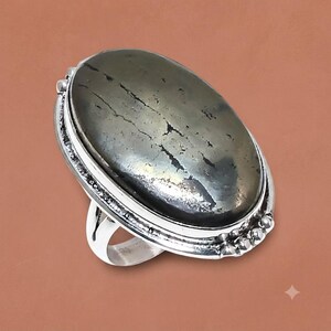 May include: A silver ring featuring a large, oval-shaped, metallic gray pyrite gemstone with natural surface fissures. The stone is set in a polished silver bezel with a decorative beaded border along the outer edge of the setting.
