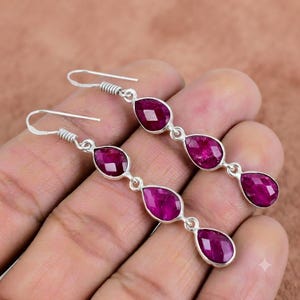 May include: A pair of long drop earrings featuring three faceted, pear-shaped magenta-coloured stones set in silver-toned metal. Each earring hangs from a simple silver hook, with the stones arranged vertically in a graduated, dangling design.