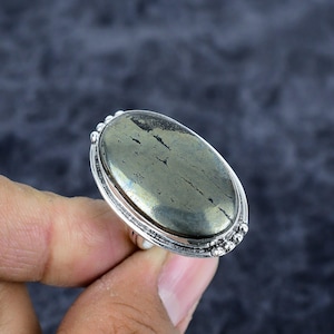 Puede incluir: Un anillo de plata con una piedra de pirita ovalada e iridiscente. La piedra tiene un brillo metálico con vetas naturales. El engaste del anillo tiene un borde decorativo con cuentas. El anillo está sostenido por una mano.
