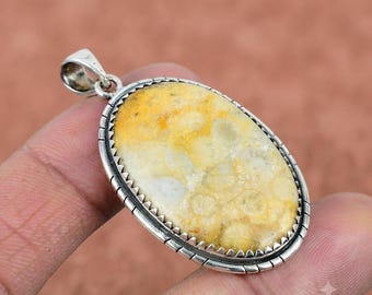 May include: An oval pendant featuring a polished, cream and yellow patterned stone set in a silver frame with a serrated edge. The pendant includes a silver bail at the top for attachment to a chain, showcasing a smooth, rounded surface texture.