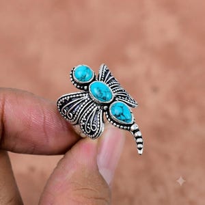 May include: A silver ring shaped like a dragonfly, featuring three turquoise stones with dark veining. The wings and body are crafted with intricate metalwork, including beaded edges and scroll patterns, creating a detailed, textured appearance.