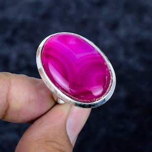 Puede incluir: Un anillo de plata con una gema rosa pulida, de forma ovalada y con un patrón de bandas. La gema está engastada en un bisel de plata, creando un contraste llamativo. El anillo es sostenido por los dedos de una persona sobre un fondo oscuro.