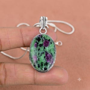Op de afbeelding: Een ovaalvormige hanger met een gepolijste zoisietsteen met levendige groene, diep magenta en zwarte spikkels. De steen is gezet in een zilverkleurige metalen rand met een decoratief oogje, bevestigd aan een dunne zilverkleurige ketting.