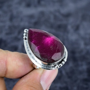 May include: A silver ring featuring a large, faceted, teardrop-shaped gemstone in a deep magenta hue. The gemstone is the focal point, set in a detailed silver band with small beaded accents. The ring's design is elegant and eye-catching.