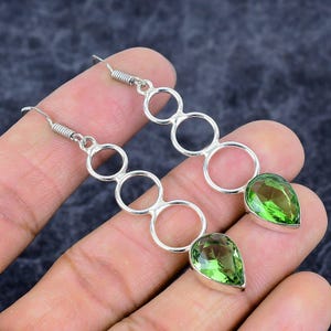 May include: A pair of silver earrings featuring a series of connected circles and a teardrop-shaped, light green gemstone. The earrings are held up by a hand against a dark background. The gemstones have a faceted cut.