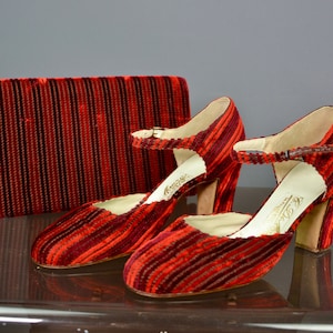 Puede incluir: Un par de zapatos de tacón de terciopelo rojo y negro a rayas con un bolso de mano a juego. Los zapatos tienen un cierre de hebilla y el bolso de mano tiene un cierre de solapa.
