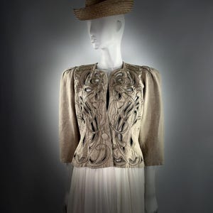 Könnte beinhalten: Eine beige Leinenjacke mit einem filigranen, durchbrochenen Design. Die Jacke hat eine figurbetonte Silhouette und einen leicht bauschigen Ärmel. Die Jacke wird mit einem weißen Rock kombiniert.