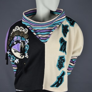 Peut inclure: Un pull noir et blanc avec un col rayé violet et bleu. Le pull a un grand motif brodé sur l'épaule gauche et des caractères japonais sur le côté droit.