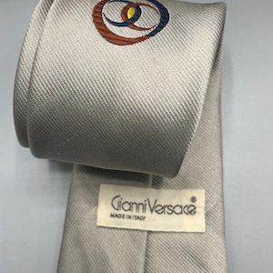 Puede incluir: Una corbata de seda plateada con un logotipo azul marino, naranja y amarillo que dice "Gianni Versace" y "Made in Italy".