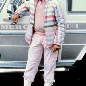 May include: A man wearing a pink and white striped jacket, white pants, and white boots stands next to a silver car with the word "MERC" visible on the side. The car has a 4x4 emblem on the back.