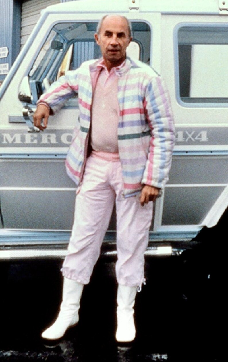 May include: A man wearing a pink and white striped jacket, white pants, and white boots stands next to a silver car with the word "MERC" visible on the side. The car has a 4x4 emblem on the back.