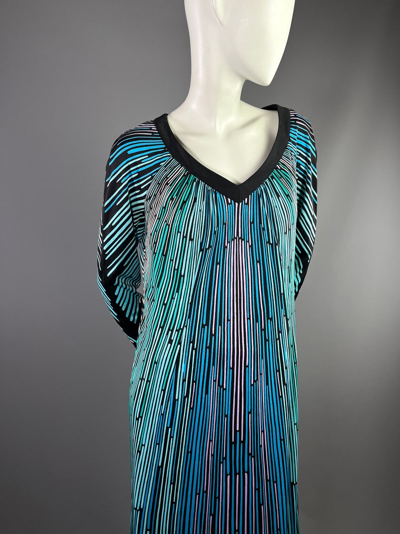 Peut inclure: Une robe maxi noire et bleue &agrave; imprim&eacute; g&eacute;om&eacute;trique avec un col en V. La robe pr&eacute;sente un motif g&eacute;om&eacute;trique de lignes verticales dans des tons de bleu, vert et rose.