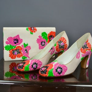 Puede incluir: Un par de zapatos blancos de tacón alto con estampado floral de flores rosas, rojas y verdes. También se muestra un bolso de mano blanco a juego con el mismo estampado floral.