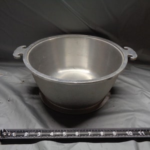 May include: A silver-colored metal cooking pot with two handles. The pot is round with a wide opening and sits on a light-colored surface. A ruler is in the foreground.