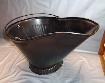 May include: A black metal coal scuttle with a wide, angled opening and a rounded base. The bucket features a ribbed texture along the sides and a thin, arched metal handle attached to the top rim, designed for carrying or scooping solid materials.