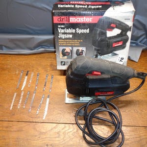 May include: A black and red Drill Master variable speed jigsaw with a box and several saw blades. The box displays the product name and a picture of the jigsaw. The jigsaw has a black power cord.