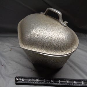 May include: A silver-colored, textured metal lunchbox with a handle on top. The lunchbox has a rounded, domed lid and a rectangular base. The lunchbox is sitting on a gray surface.
