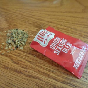 May include: A red packet of "Original Seasoning Blend" with the brand name "House-Autry" and a small pile of seasoning granules on a wooden surface. The packet is salt-free and contains 0.03 oz of seasoning.
