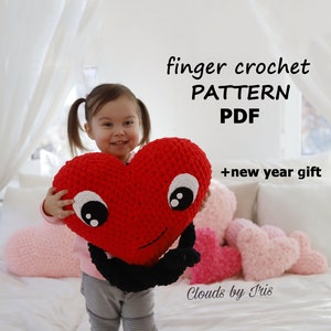 Könnte beinhalten: Ein Kind hält ein rotes, gehäkeltes Herzförmiges Kissen mit schwarzen Augen und einem lächelnden Mund. Das Kissen ist umgeben von anderen gehäkelten Herzförmigen Kissen in Rosa und Weiß. Der Text "finger crochet PATTERN PDF + new year gift" wird über den Kissen angezeigt. Der Text "Clouds by Iris" wird unter den Kissen angezeigt.
