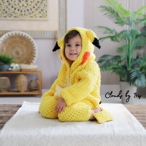 May include: A child wearing a yellow crocheted Pikachu costume with black and red accents. The costume features a hood with ears and a tail. The child is sitting on a white blanket.
