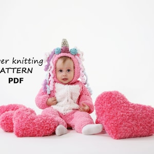 Puede incluir: Un disfraz de unicornio rosa de tejido de dedos para un bebé. El disfraz tiene una barriga blanca, una melena rosa y un cuerno azul y blanco. El bebé está sentado sobre un fondo blanco con almohadas en forma de corazón rosa.