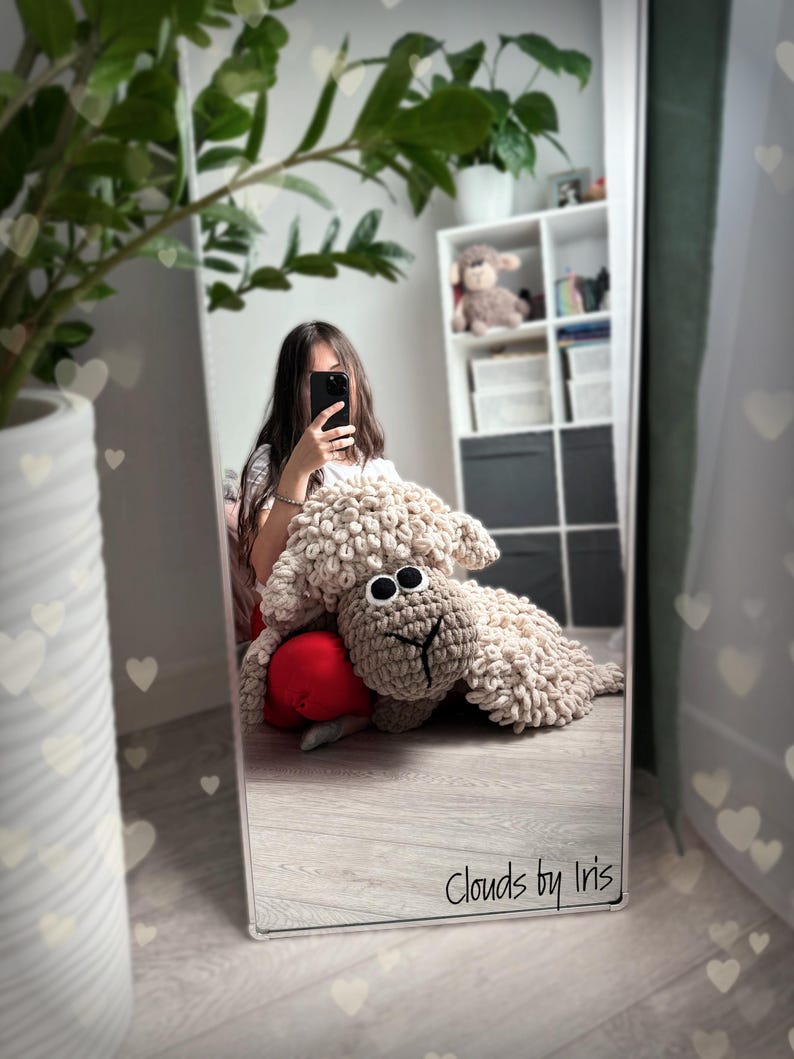 May include: A crocheted sheep plush toy with brown eyes and a light brown nose. The sheep is lying on a wooden floor in front of a mirror. The text "Clouds by Iris" is visible in the reflection.