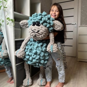 Może przedstawiać: Młoda dziewczyna trzyma w ramionach dużą pluszową owcę wykonaną na szydełku. Owca jest jasnoniebieska i szara z przyjaznym uśmiechem. Dziewczyna uśmiecha się i ma na sobie dżinsy. Widać napis "Clouds by Iris".