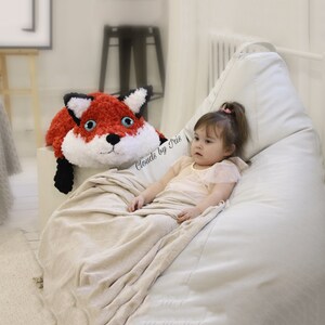 May include: A young child sits in a beanbag chair with a red and white crocheted fox plush toy. The child is wearing a pink shirt and the beanbag chair is white. The text "Clouds by Iris" is visible in the image.