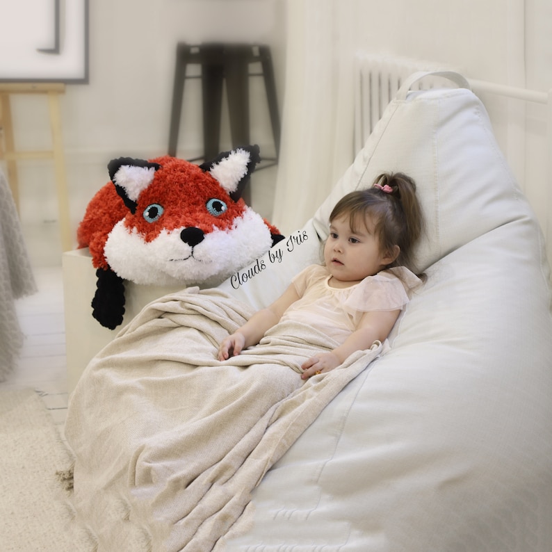 May include: A young child sits in a beanbag chair with a red and white crocheted fox plush toy. The child is wearing a pink shirt and the beanbag chair is white. The text "Clouds by Iris" is visible in the image.