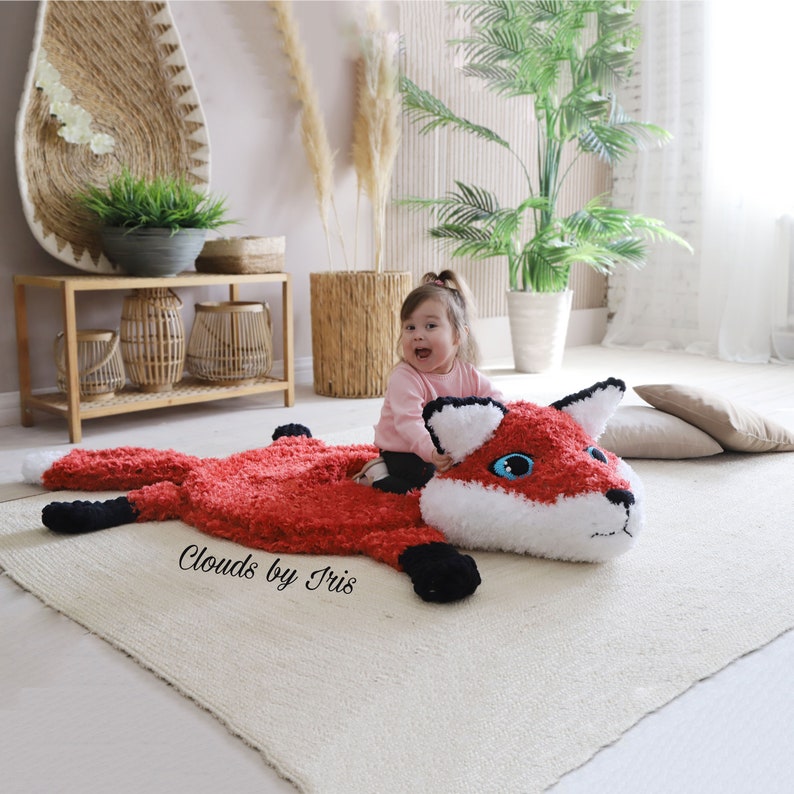 May include: A red and white crocheted fox rug with black accents. A young child is sitting on the rug, smiling. The rug has the text "Clouds by Iris" on it.