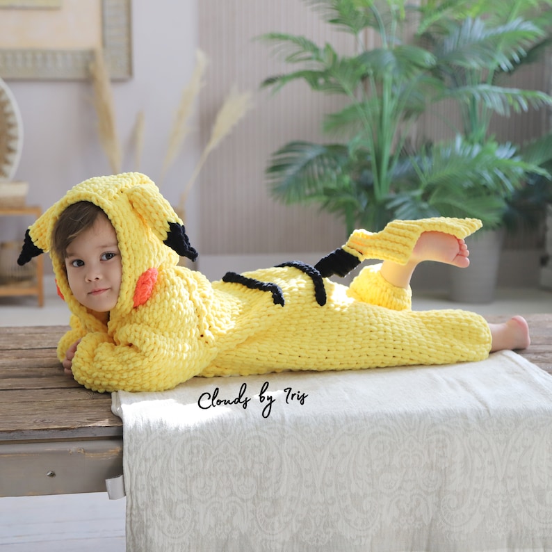 May include: A yellow knitted baby costume with black and red details, resembling a popular cartoon character. The costume features a hood with pointed ears and a tail. The baby is lying on a wooden surface with a white blanket.