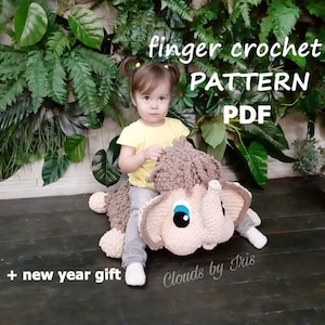 May include: A young child sits on a crocheted mammoth toy. The toy is brown and beige with blue eyes. The text "finger crochet PATTERN PDF + new year gift Clouds by Iris" is on the image.