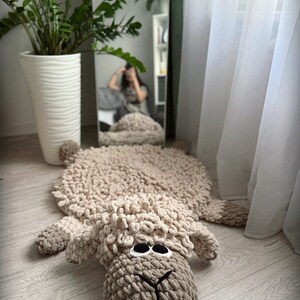 May include: A beige crocheted sheep rug with large black eyes. The rug is lying on a light wood floor. The text "Clouds by Iris" is in the lower left corner of the image.
