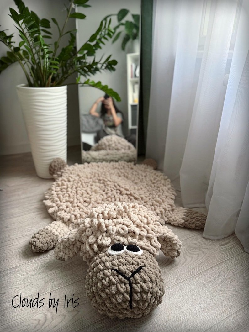 May include: A beige crocheted sheep rug with large black eyes. The rug is lying on a light wood floor. The text "Clouds by Iris" is in the lower left corner of the image.