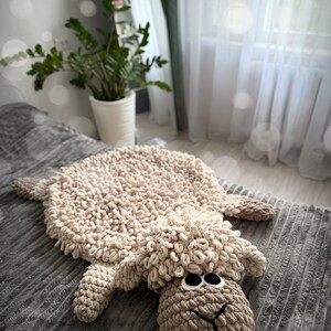May include: A beige crocheted sheep rug with a fluffy texture. The rug is lying on a gray textured surface. The sheep has large black eyes and a brown nose. The text "Clouds by Iris" is visible in the bottom left corner of the image.