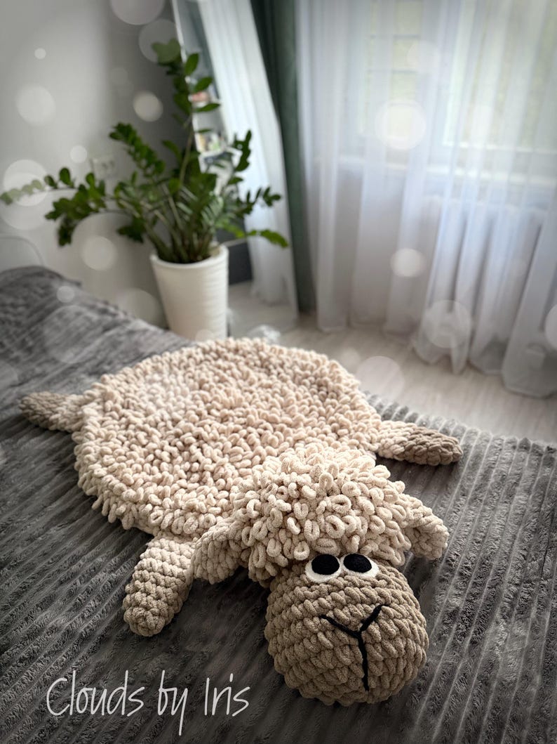 May include: A beige crocheted sheep rug with a fluffy texture. The rug is lying on a gray textured surface. The sheep has large black eyes and a brown nose. The text "Clouds by Iris" is visible in the bottom left corner of the image.