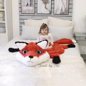 May include: A red and white crocheted fox blanket with black accents. The blanket is on a bed with a young child sitting on it. The blanket has a large fox head with blue eyes and a black nose. The text "Clouds by Iris" is visible in the bottom right corner of the image.