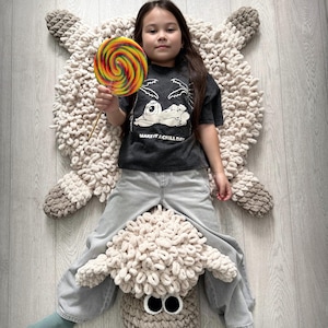 May include: A child sits on a large, cream-colored crocheted sheep rug. The rug has a brown face with large eyes and a black nose. The child is wearing a grey t-shirt with the text "Make it a chill day" and blue jeans. The child is holding a colorful lollipop.