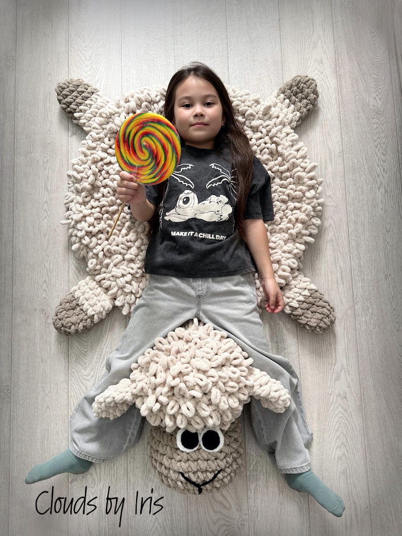May include: A child sits on a large, cream-colored crocheted sheep rug. The rug has a brown face with large eyes and a black nose. The child is wearing a grey t-shirt with the text "Make it a chill day" and blue jeans. The child is holding a colorful lollipop.
