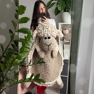 May include: A beige crocheted sheep rug with a fluffy texture. The rug has a large head with black eyes and a brown nose. The rug is being held by a person in a white shirt and red pants. The text "Clouds by Iris" is visible in the bottom right corner of the image.